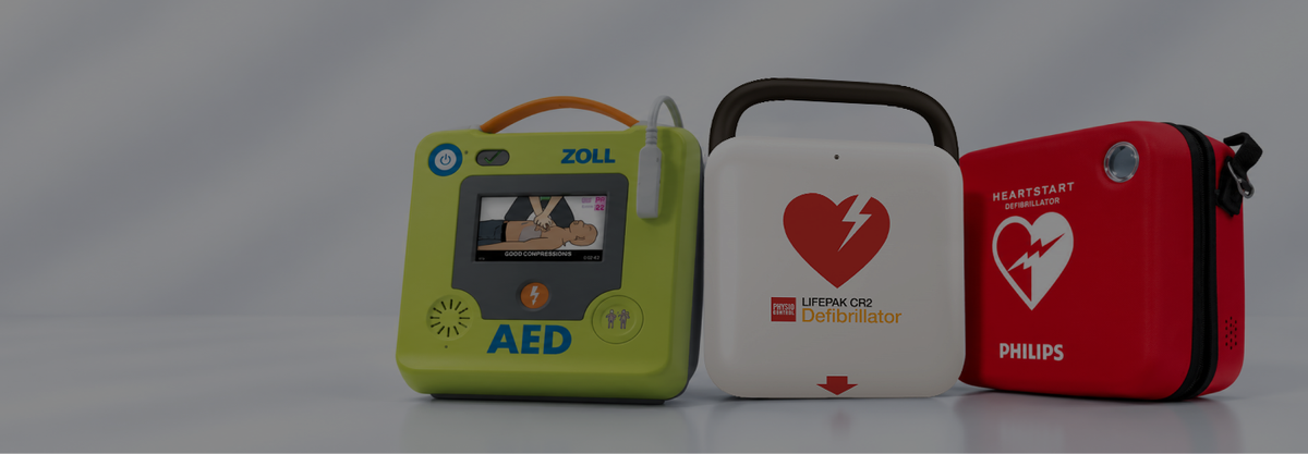 Three AEDs and a red carrying case shown side by side on a gray studio background: a lime green ZOLL AED, a white Lifepak CR2, and a red Philips HeartStart case.