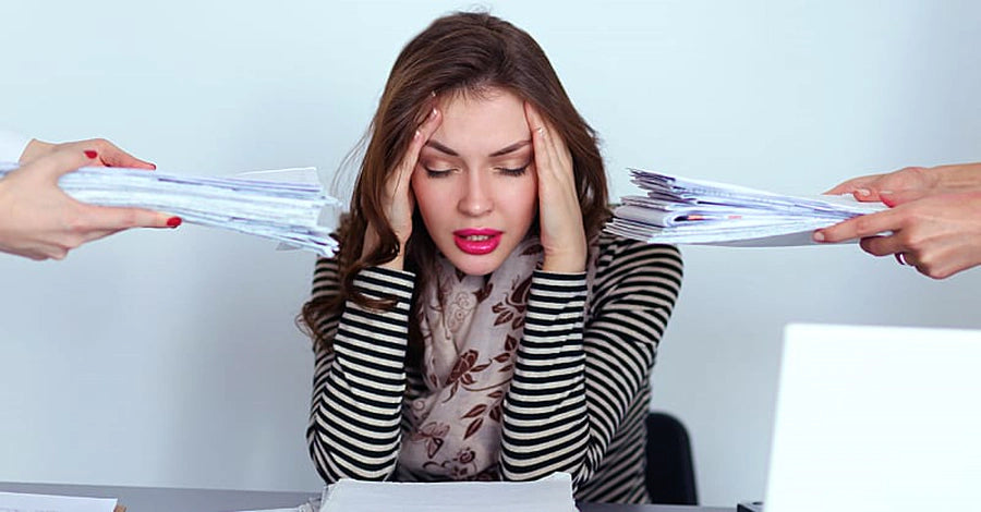 Stressed woman holding her head at a desk while two hands hold out stacks of papers