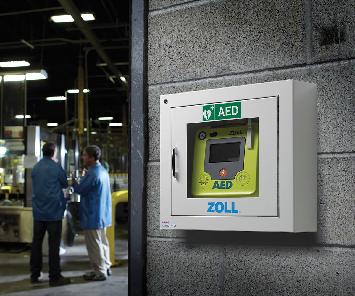 Two men stand in a factory with a ZOLL AED 3 hanging in a cabinet on a concrete block wall
