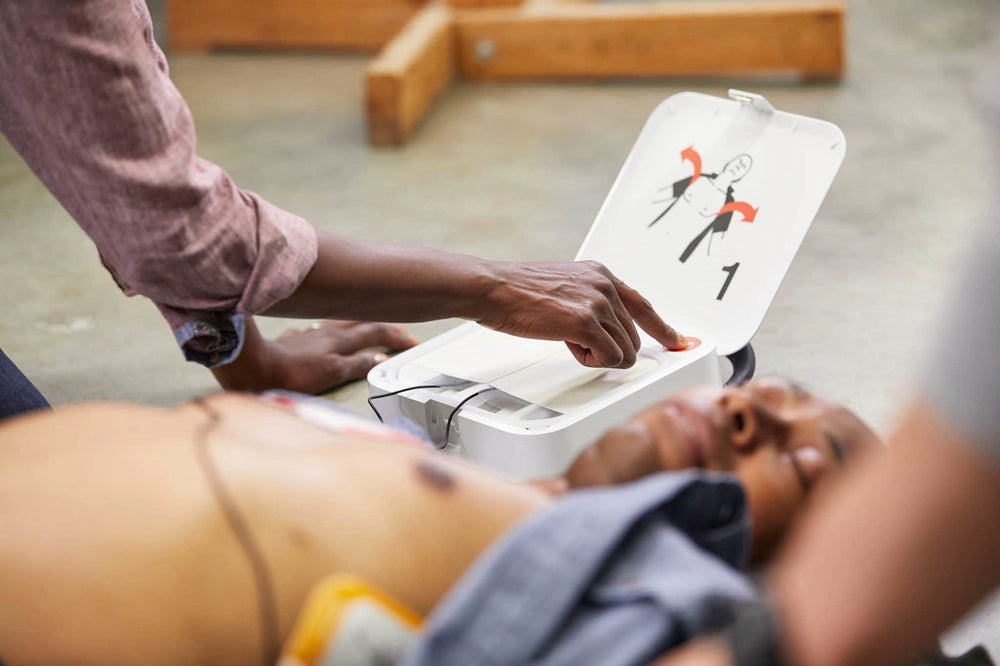 Person's hand pressing the shock button on an open AED training device beside a manikin