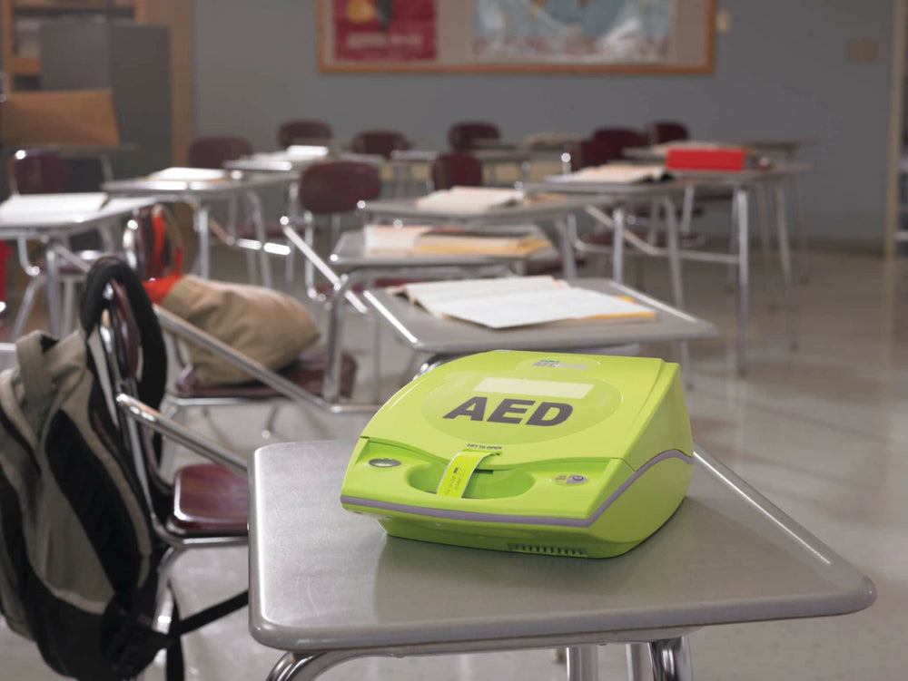 Green AED device sitting on a classroom desk with empty desks in the background