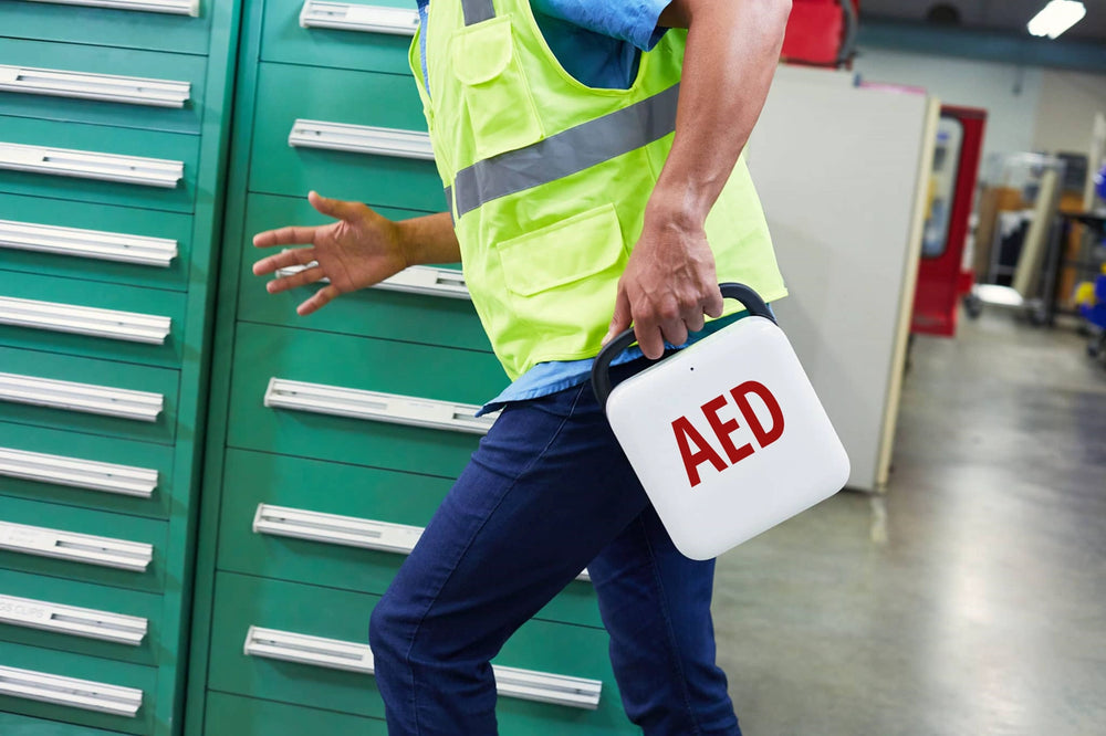 Worker in a safety vest carrying a white AED case marked AED through a warehouse