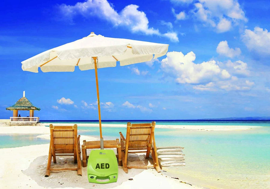 Green AED case placed on a sandy beach beneath a white umbrella between two lounge chairs