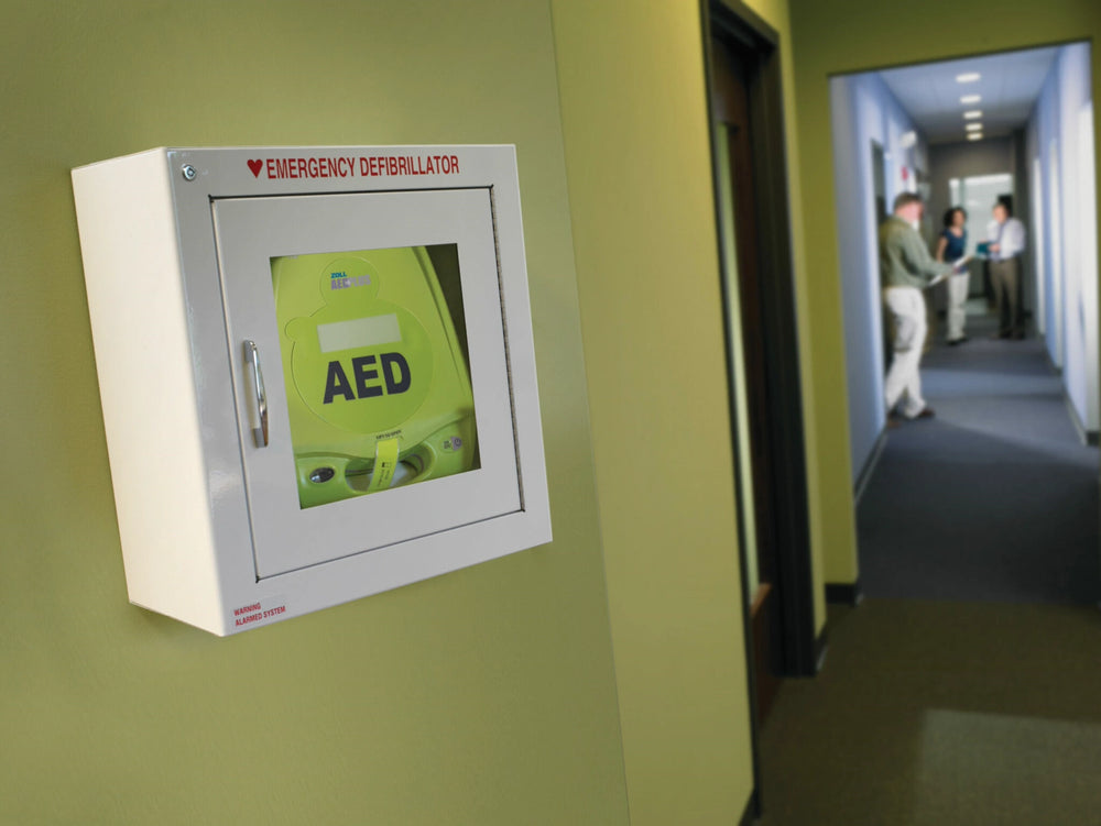 Wall-mounted green AED emergency defibrillator cabinet in an office hallway with people talking in the background
