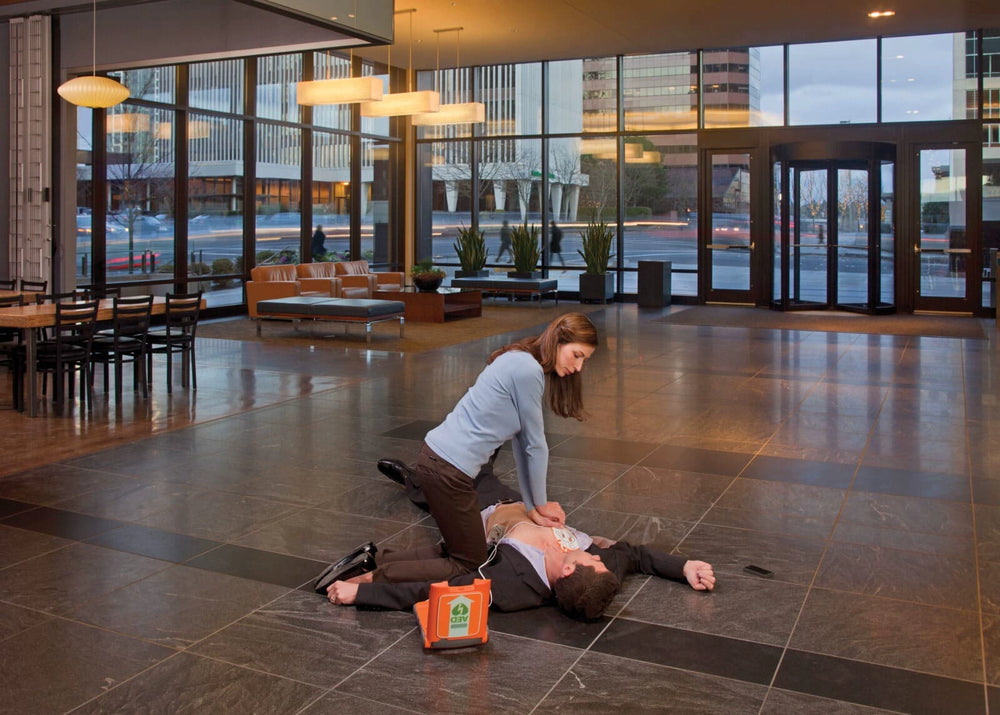 Woman performing chest compressions on a man lying on the floor of an office lobby, with an AED case nearby