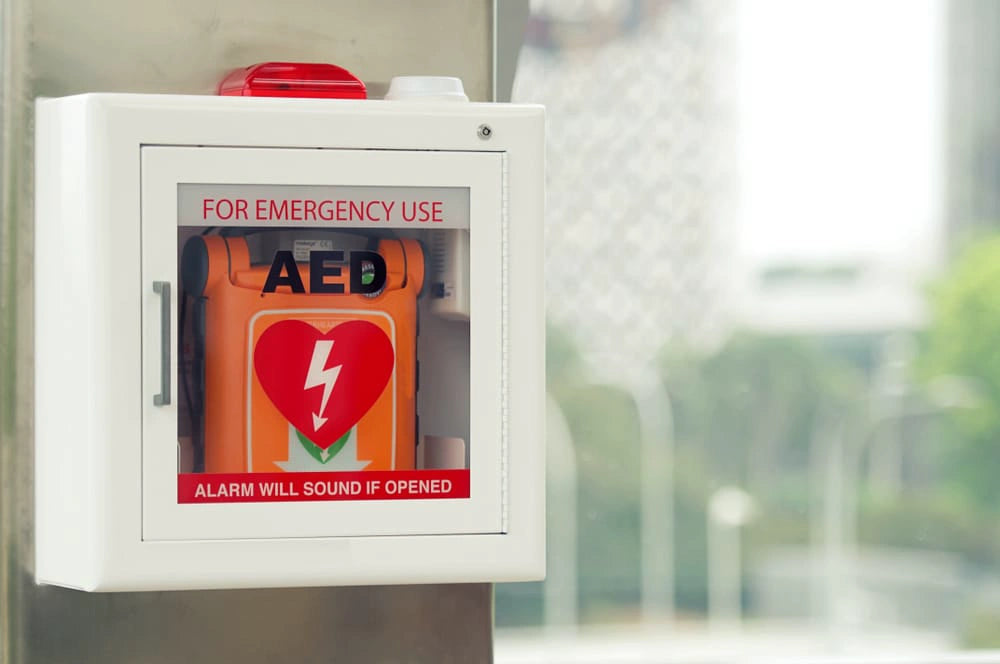 Wall-mounted white cabinet with an orange AED device visible behind glass labeled For Emergency Use