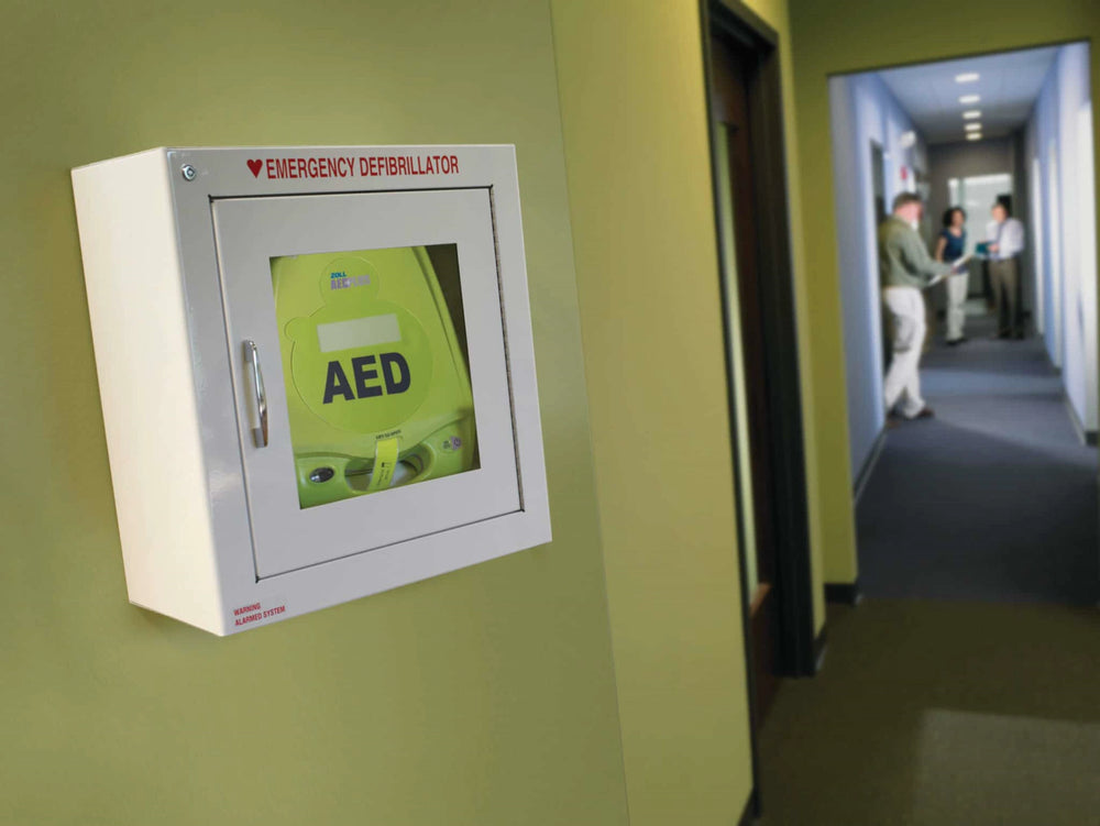 Wall-mounted green emergency defibrillator cabinet in an office hallway with people talking in the background