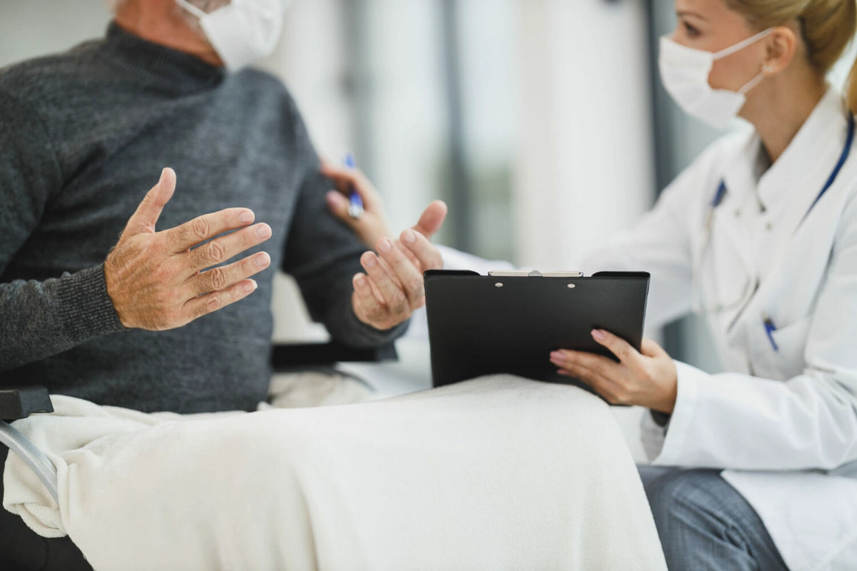 Elderly man wearing a face mask gesturing while talking with a masked doctor holding a clipboard